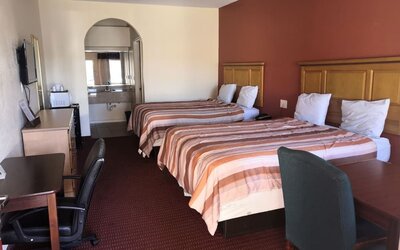 Guest room with two beds, striped orange and brown bedspreads, wooden headboards, desks and office chairs, television, and bathroom vanity visible through an arched opening at Scottish Inn & Suites.