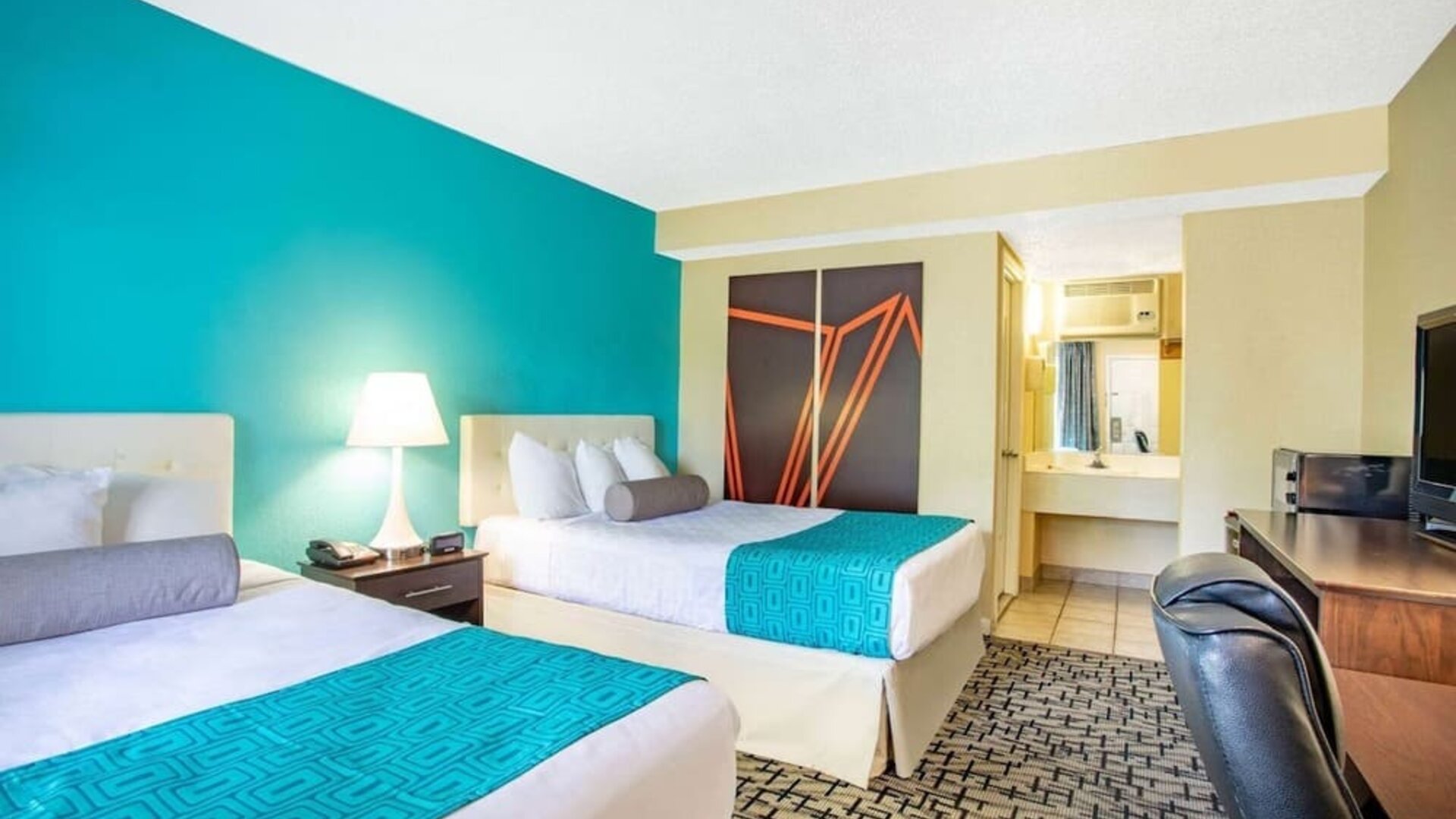 Guest room with two beds featuring white bedding and turquoise patterned runners, tufted headboards, bedside lamp, desk and chair, television, and bathroom vanity visible through an open doorway at Howard Johnson Express Inn - Rocky Hill.
