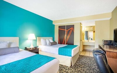Guest room with two beds featuring white bedding and turquoise patterned runners, tufted headboards, bedside lamp, desk and chair, television, and bathroom vanity visible through an open doorway at Howard Johnson Express Inn - Rocky Hill.