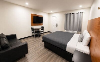Bright contemporary king room featuring a large bed with gray and white bedding, oversized wood-paneled headboard, black loveseat, wall-mounted flat-screen TV, small dining table with two chairs, air-conditioning unit beneath gray curtains, recessed light