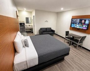 Spacious modern king room with a large bed dressed in gray and white linens, wood-paneled headboard, black loveseat, wall-mounted flat-screen TV, dining table with two chairs, microwave, mini refrigerator, vanity area, recessed lighting, and wood-look flo
