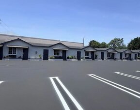 Daytime view of the Scenic Motel parking area with a clearly marked accessible parking space in the foreground and rows of single-story guest rooms with individual entrances, windows, small awnings, landscaped shrubs, and parking spaces extending into the