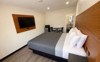 Bright modern guest room with a king-size bed dressed in gray and white bedding, a large wood-paneled headboard, wall-mounted flat-screen TV, small dining table with two chairs, microwave, mini refrigerator, wood-look flooring, and an open doorway leading