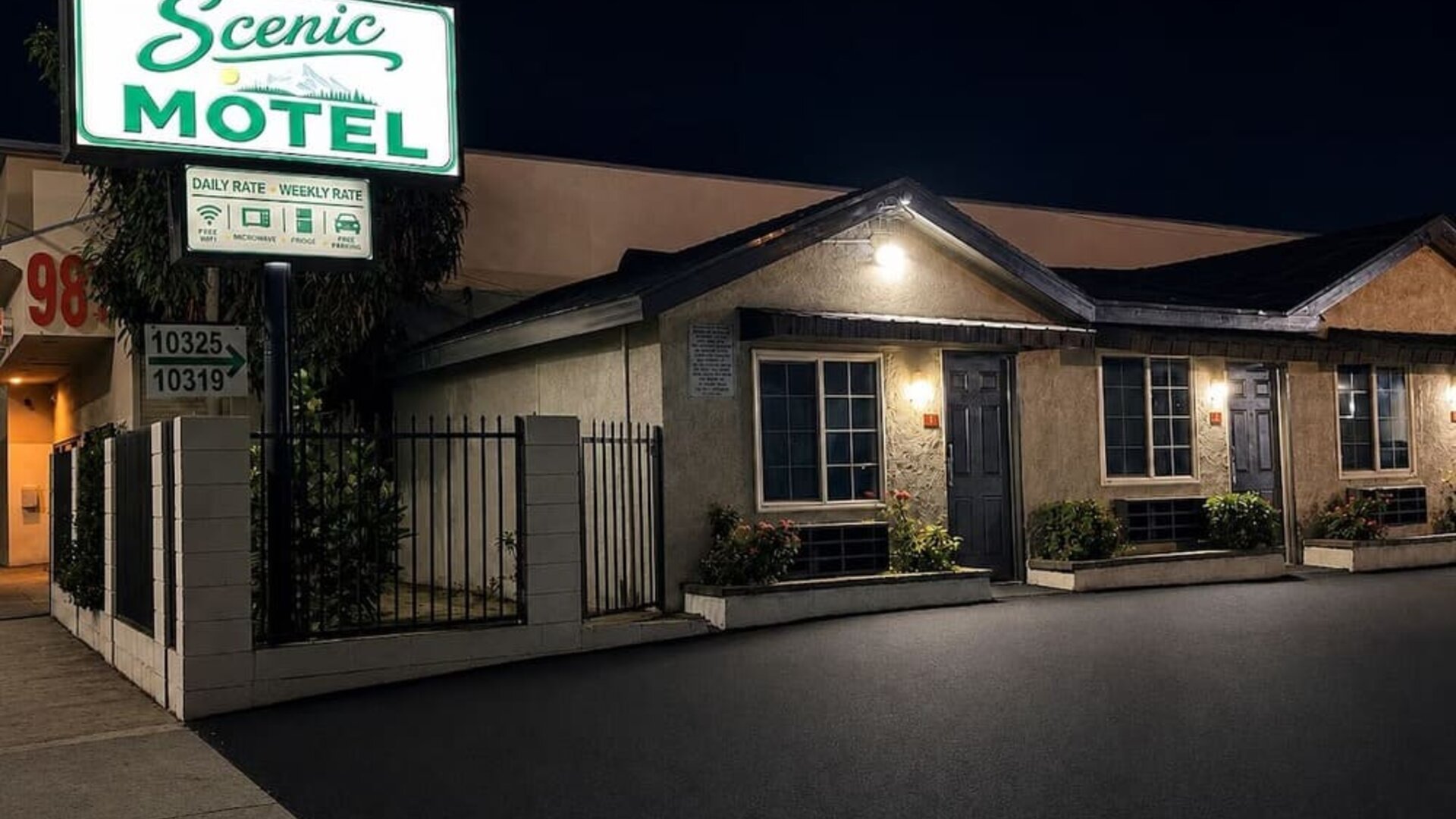Nighttime exterior view of Scenic Motel with a large illuminated green-and-white roadside sign above the single-story property, where individual guest-room entrances, windows, exterior lights, landscaped planters, and the paved parking area are visible.