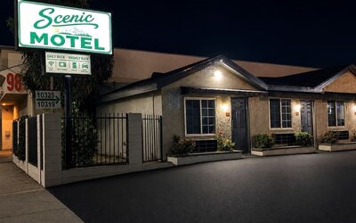 Nighttime exterior view of Scenic Motel with a large illuminated green-and-white roadside sign above the single-story property, where individual guest-room entrances, windows, exterior lights, landscaped planters, and the paved parking area are visible.