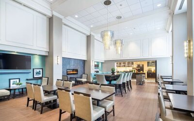 Bright hotel dining area with modern tables and chairs, high-top communal seating, contemporary pendant lights, a fireplace, and a serving area in the background.