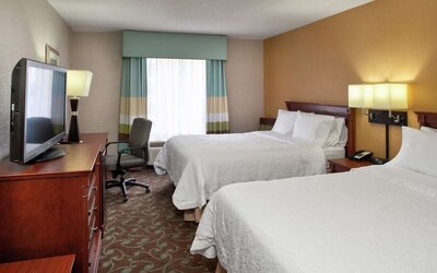 Comfortable hotel day room with two beds dressed in white linens, work desk and chair, flat-screen TV, wooden furnishings, and a large window with striped curtains.