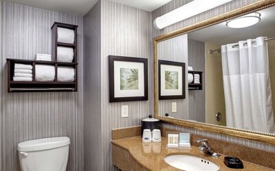Hotel guest bathroom with a granite vanity, sink, large framed mirror, shower with white curtain, toilet, neatly arranged towels, and complimentary toiletries.