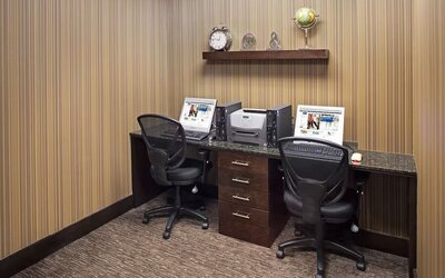 Hotel business center with two computer workstations, office chairs, a printer, granite countertop, dark wood cabinetry, and a decorative wall shelf.