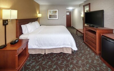 Spacious hotel guest room featuring a king-size bed with white linens, bedside lighting, wooden furnishings, flat-screen TV, dresser, desk, and mini refrigerator.
