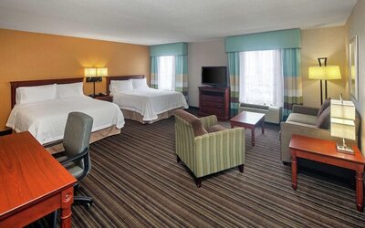 Spacious hotel guest room with two queen beds, white linens, seating area with sofa and armchairs, work desk, flat-screen TV, and two large windows.