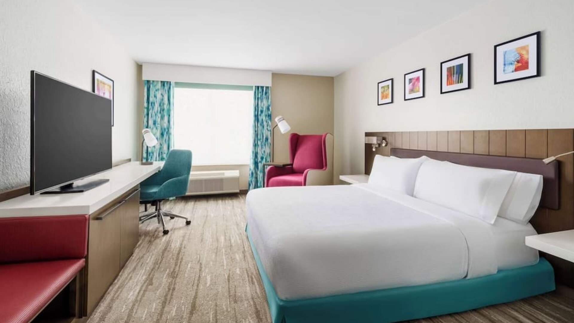 Spacious hotel room with a king bed, turquoise bed base, large TV, work desk, teal office chair, red accent seating and colorful framed artwork.