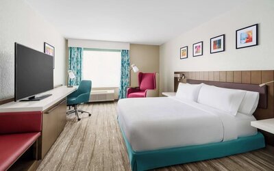 Spacious hotel room with a king bed, turquoise bed base, large TV, work desk, teal office chair, red accent seating and colorful framed artwork.