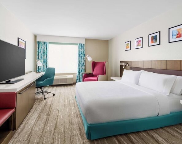 Spacious hotel room with a king bed, turquoise bed base, large TV, work desk, teal office chair, red accent seating and colorful framed artwork.