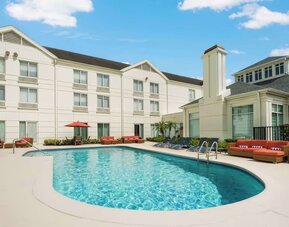 Curved outdoor hotel swimming pool with lounge chairs, orange cushioned seating, an umbrella and a bright three-story hotel building surrounding the courtyard.