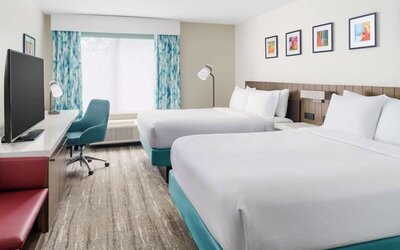 Bright modern hotel guest room with two white beds, turquoise bed bases, a large TV, work desk with teal chair, colorful artwork and patterned blue curtains.