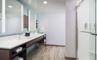 Spacious modern hotel bathroom with a long double-sink vanity, illuminated mirrors, open towel storage and a glass-enclosed walk-in shower.