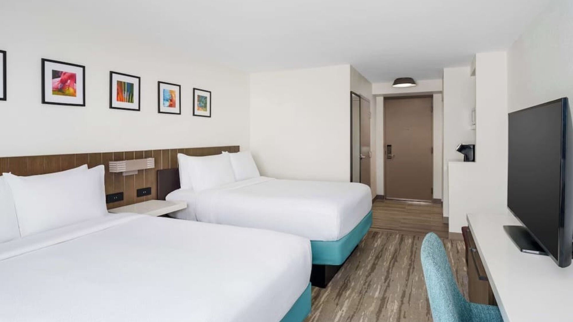 Spacious hotel guest room with two white beds on turquoise bases, colorful framed artwork, large flat-screen TV, teal desk chair and modern wood-look flooring.`
