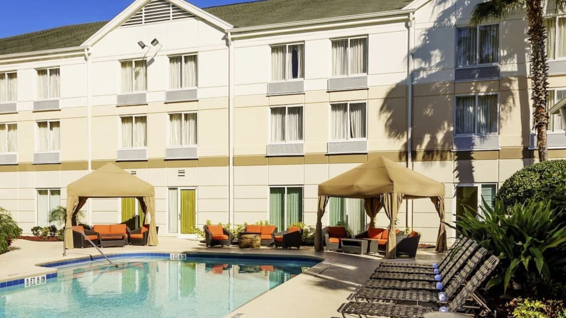 Outdoor hotel swimming pool surrounded by lounge chairs, shaded cabanas with orange seating, tropical landscaping and the hotel building.