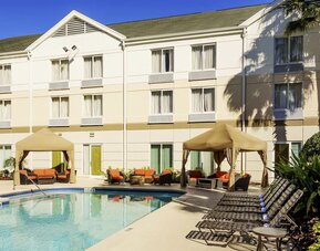 Outdoor hotel swimming pool surrounded by lounge chairs, shaded cabanas with orange seating, tropical landscaping and the hotel building.