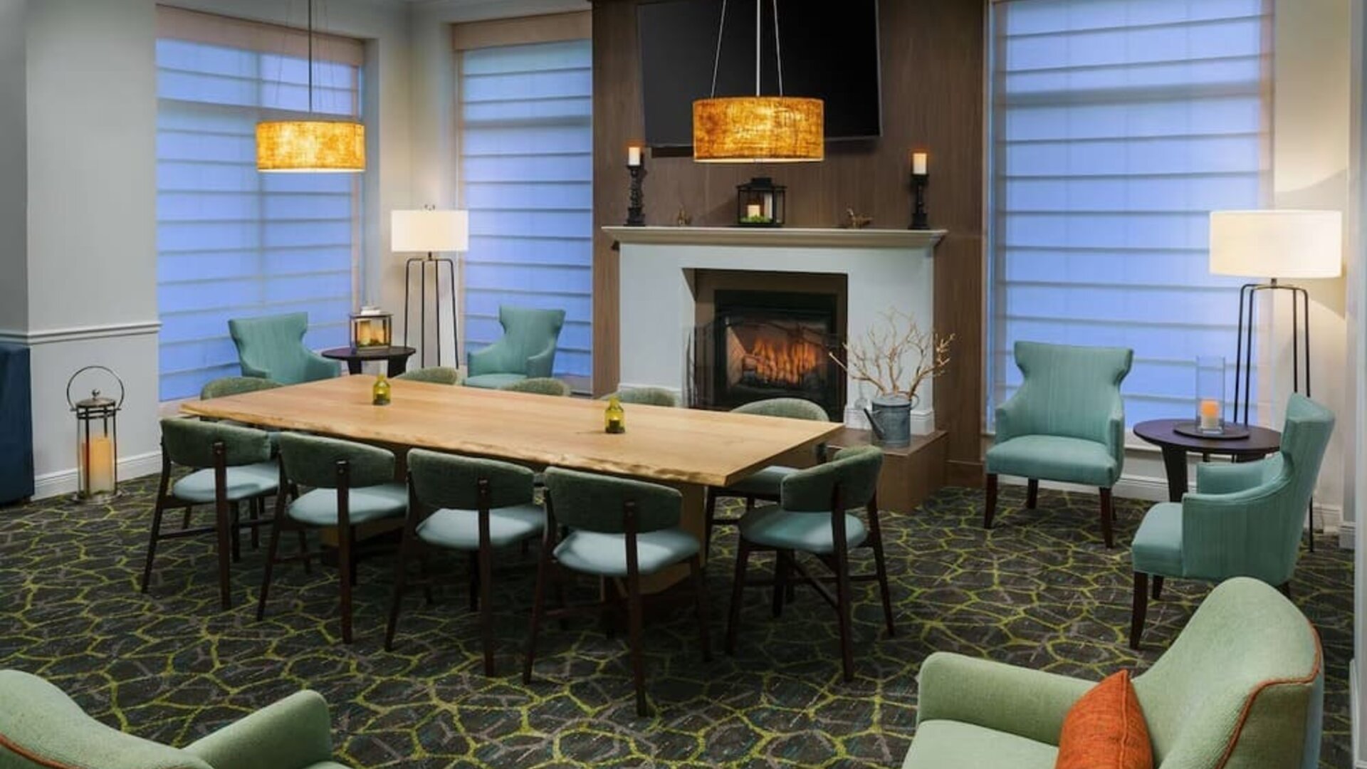 Cozy hotel lounge with a central fireplace, large communal wood table, mint-green armchairs, floor lamps and warm pendant lighting.