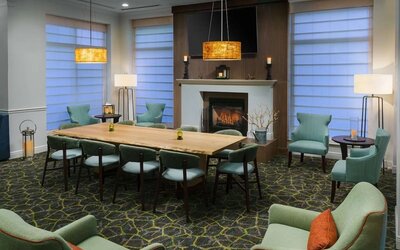 Cozy hotel lounge with a central fireplace, large communal wood table, mint-green armchairs, floor lamps and warm pendant lighting.