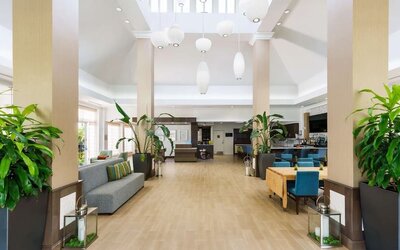 Bright, open hotel lobby with tall columns, large tropical plants, gray sofa seating, turquoise dining chairs, pendant lights and access to the bar and reception areas.