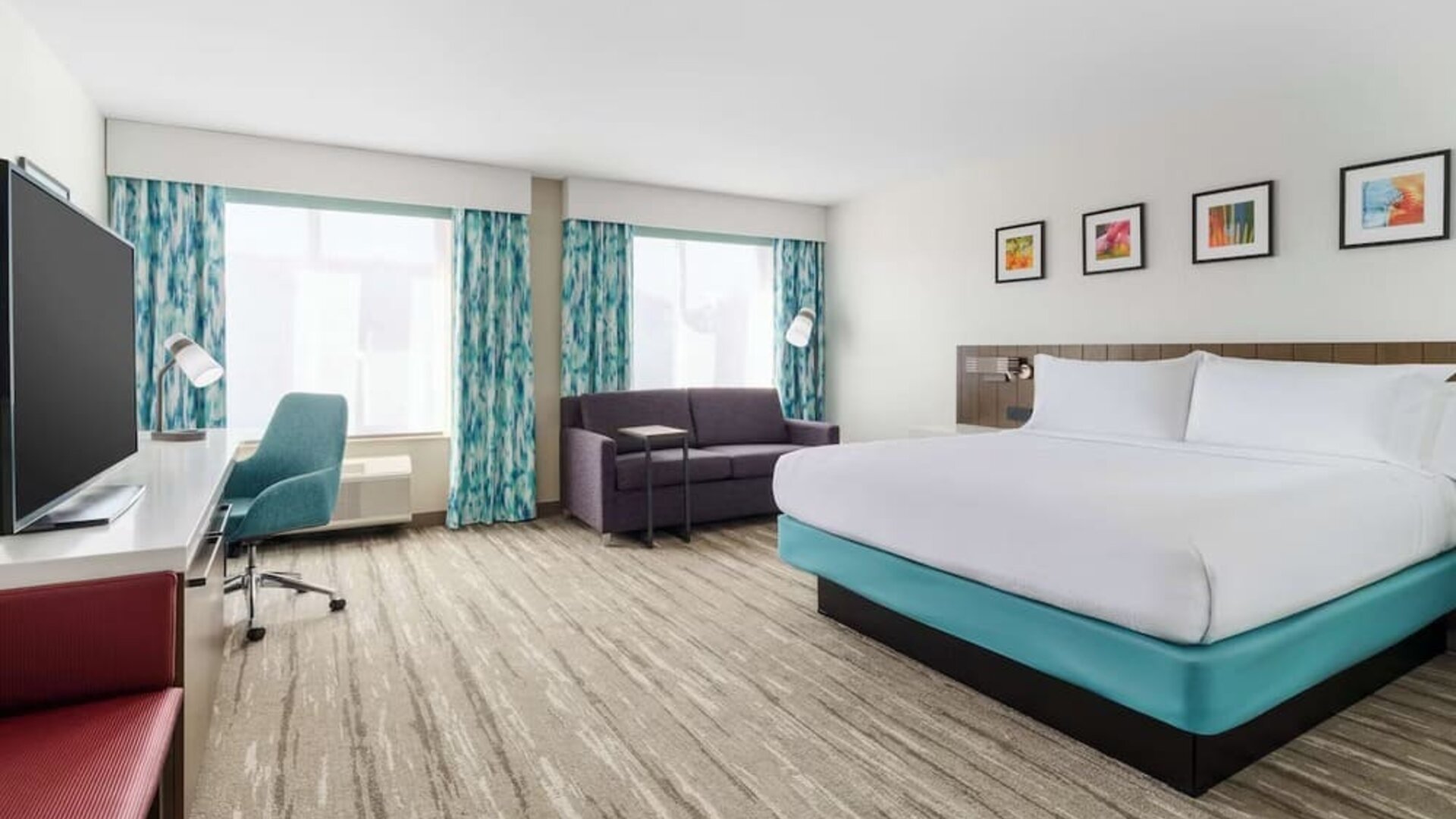 Spacious modern hotel room with a king bed, sofa, large TV, work desk, teal office chair, two large windows and colorful blue-patterned curtains.