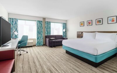 Spacious modern hotel room with a king bed, sofa, large TV, work desk, teal office chair, two large windows and colorful blue-patterned curtains.