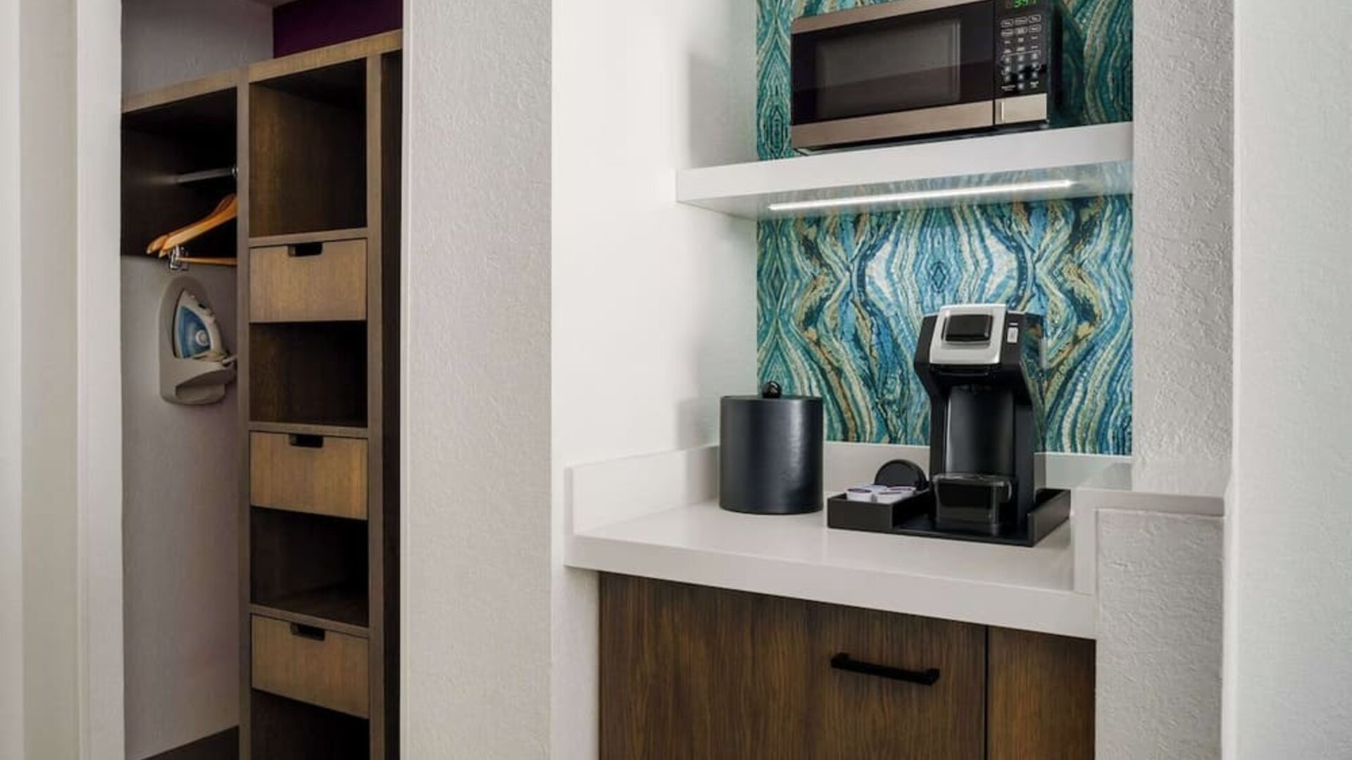 In-room coffee station with a single-serve coffee maker, microwave, ice bucket, storage cabinet, open closet, hangers and ironing board against a decorative blue accent wall.