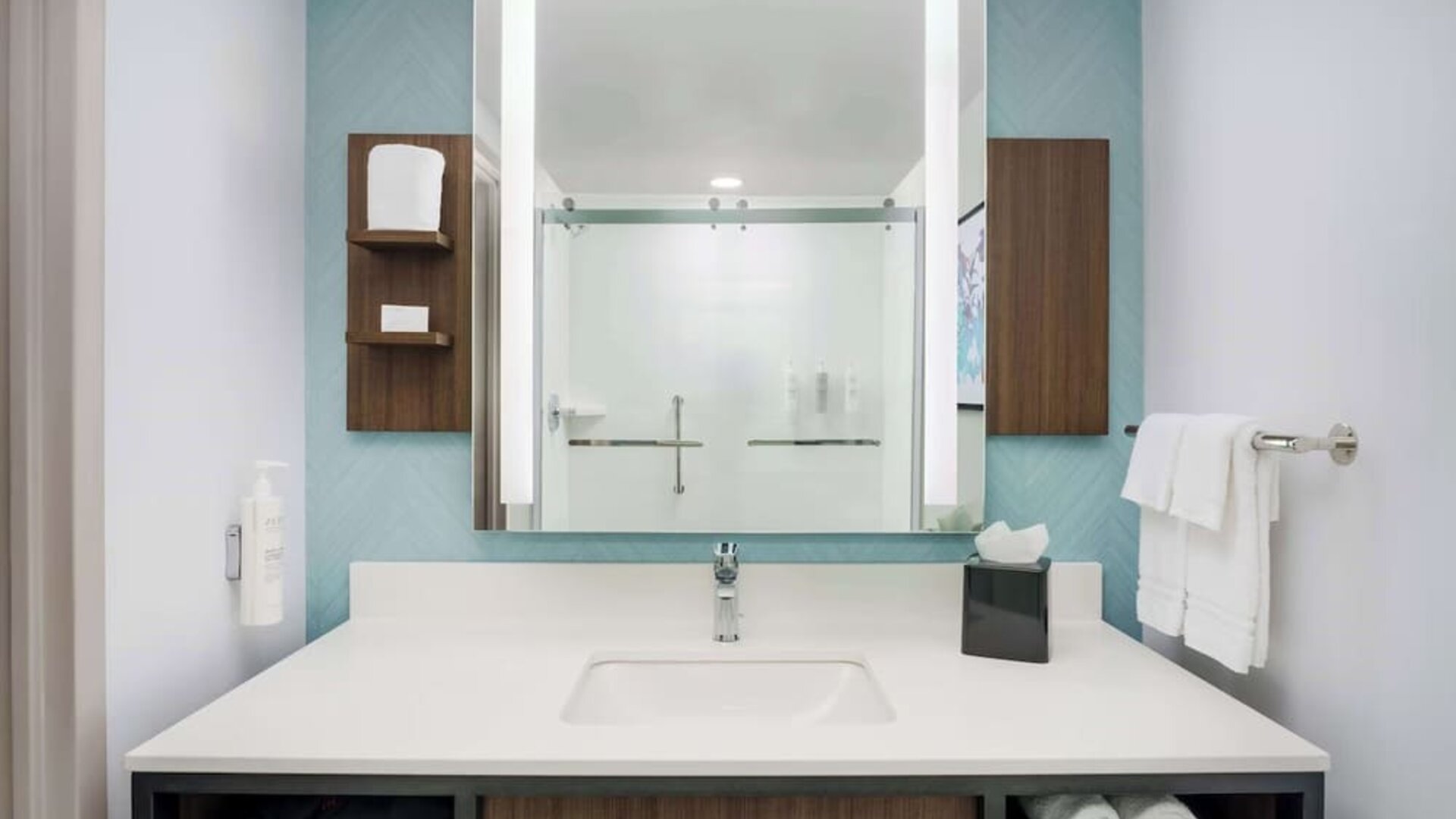 Modern hotel bathroom vanity with a white countertop and sink, large illuminated mirror, open towel storage and glass-enclosed shower reflected in the mirror.