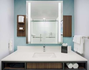 Modern hotel bathroom vanity with a white countertop and sink, large illuminated mirror, open towel storage and glass-enclosed shower reflected in the mirror.