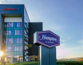Exterior of Hampton by Hilton hotel featuring a modern multi-story glass and dark-panel façade, large freestanding Hampton by Hilton sign, landscaped grounds, and blue sky.