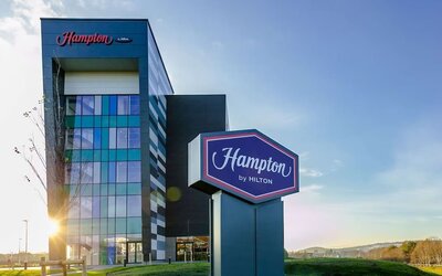 Exterior of Hampton by Hilton hotel featuring a modern multi-story glass and dark-panel façade, large freestanding Hampton by Hilton sign, landscaped grounds, and blue sky.