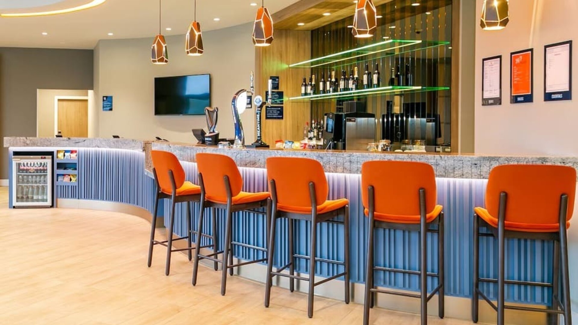 Modern Hampton by Hilton hotel bar with a long stone countertop, bright orange high-back bar stools, geometric pendant lights, beer taps, bottle displays, and illuminated shelving.