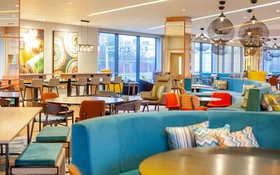 Colorful open-plan Hampton by Hilton hotel lounge with turquoise sofas, patterned cushions, assorted tables and chairs, contemporary pendant lights, large windows, and communal seating areas.