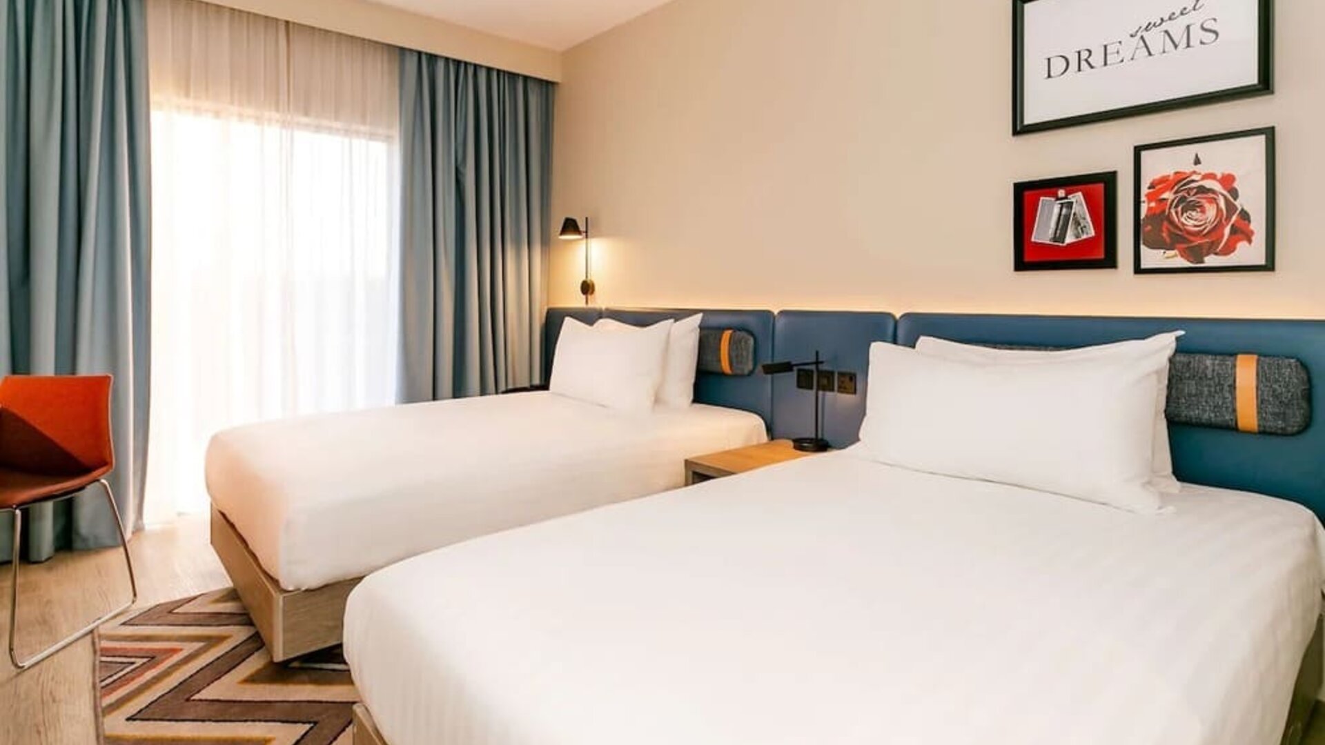 Bright contemporary hotel room with two beds, crisp white linens, blue upholstered headboard, framed wall art, floor-to-ceiling window with blue curtains, orange chair, wood flooring, and patterned rug.