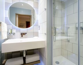 Bright contemporary hotel bathroom with a large illuminated round mirror, white rectangular sink, folded towels, glass-enclosed walk-in shower, handheld showerhead, and wall-mounted toiletries.
