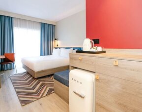 Modern hotel guest room with a king-size bed, white bedding, blue curtains, wood furnishings, red accent wall, compact SMEG refrigerator, tea and coffee station, patterned rug, and desk chair.