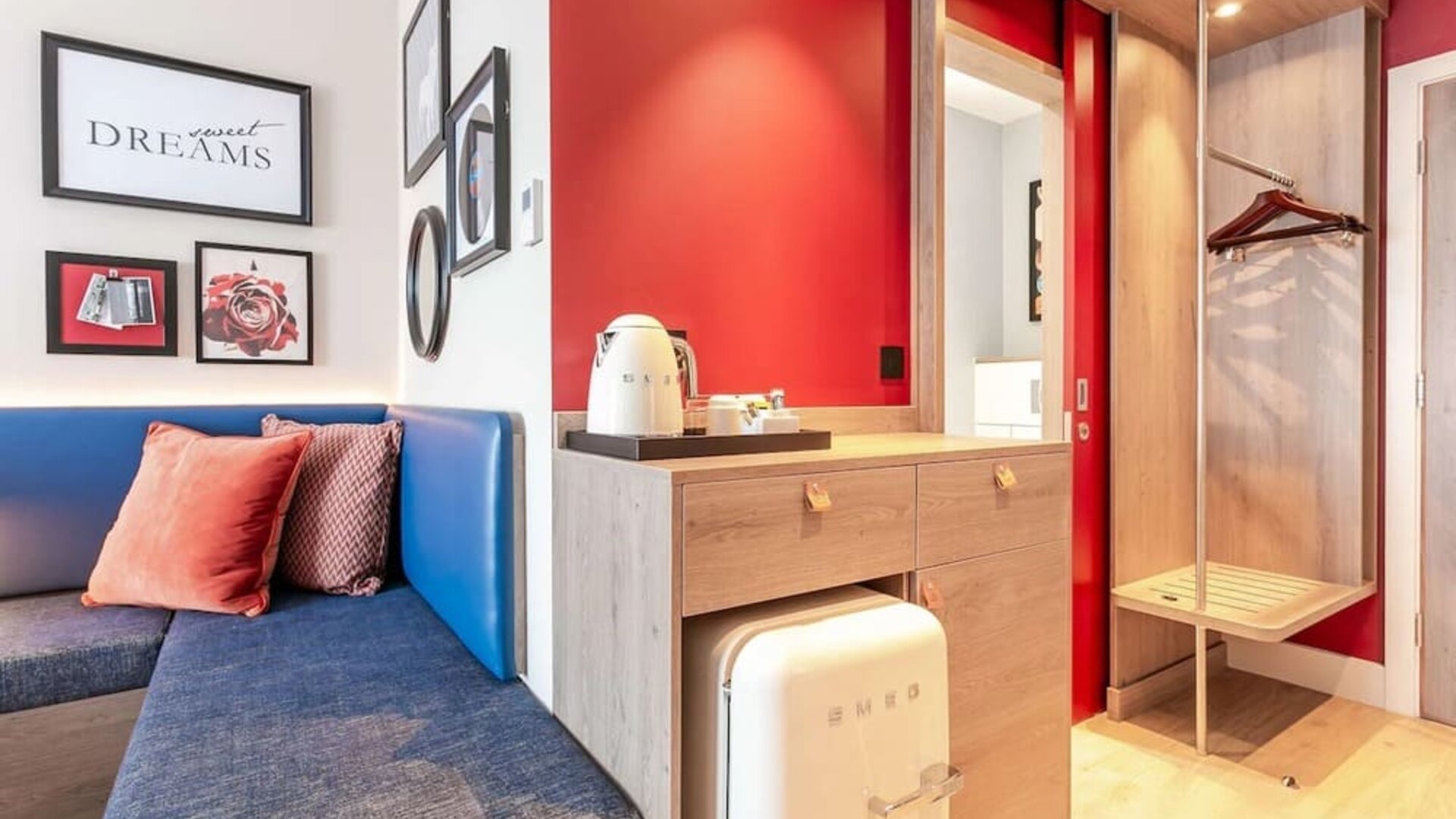 Modern hotel room amenity area with a blue upholstered bench, decorative cushions, framed artwork, tea and coffee station, compact SMEG refrigerator, open wardrobe with hangers, luggage rack, and red accent walls.