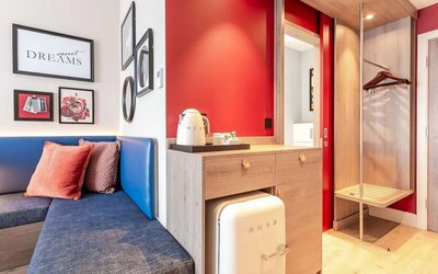 Modern hotel room amenity area with a blue upholstered bench, decorative cushions, framed artwork, tea and coffee station, compact SMEG refrigerator, open wardrobe with hangers, luggage rack, and red accent walls.