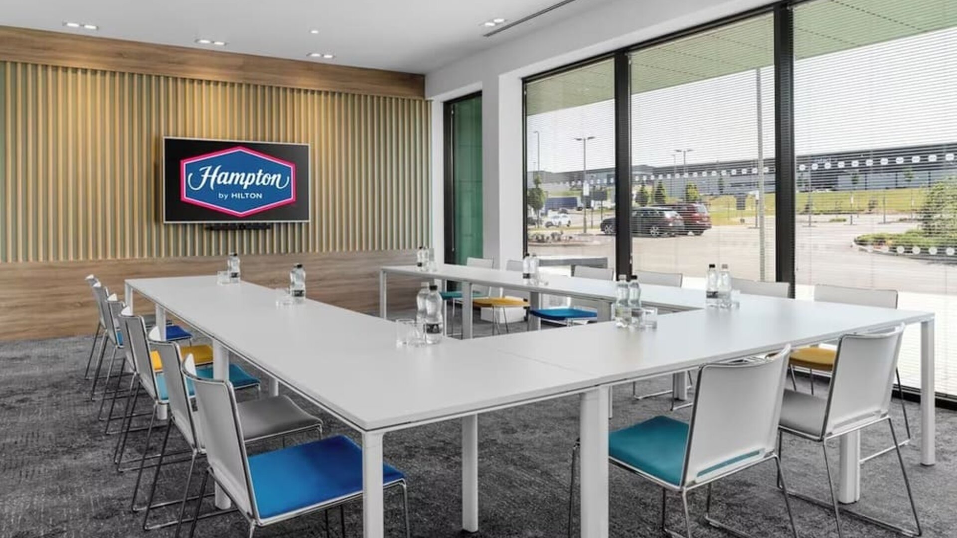 Bright Hampton by Hilton meeting room with white tables arranged in a U-shape, colorful chairs, bottled water, large windows with blinds, and a wall-mounted screen displaying the Hampton by Hilton logo.