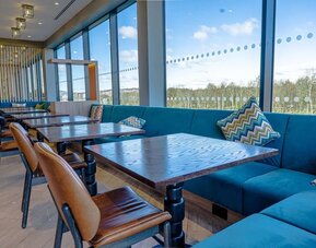 Spacious hotel dining area with large floor-to-ceiling windows, turquoise upholstered banquette seating, patterned cushions, dark wood tables, and brown leather dining chairs overlooking the surrounding landscape.