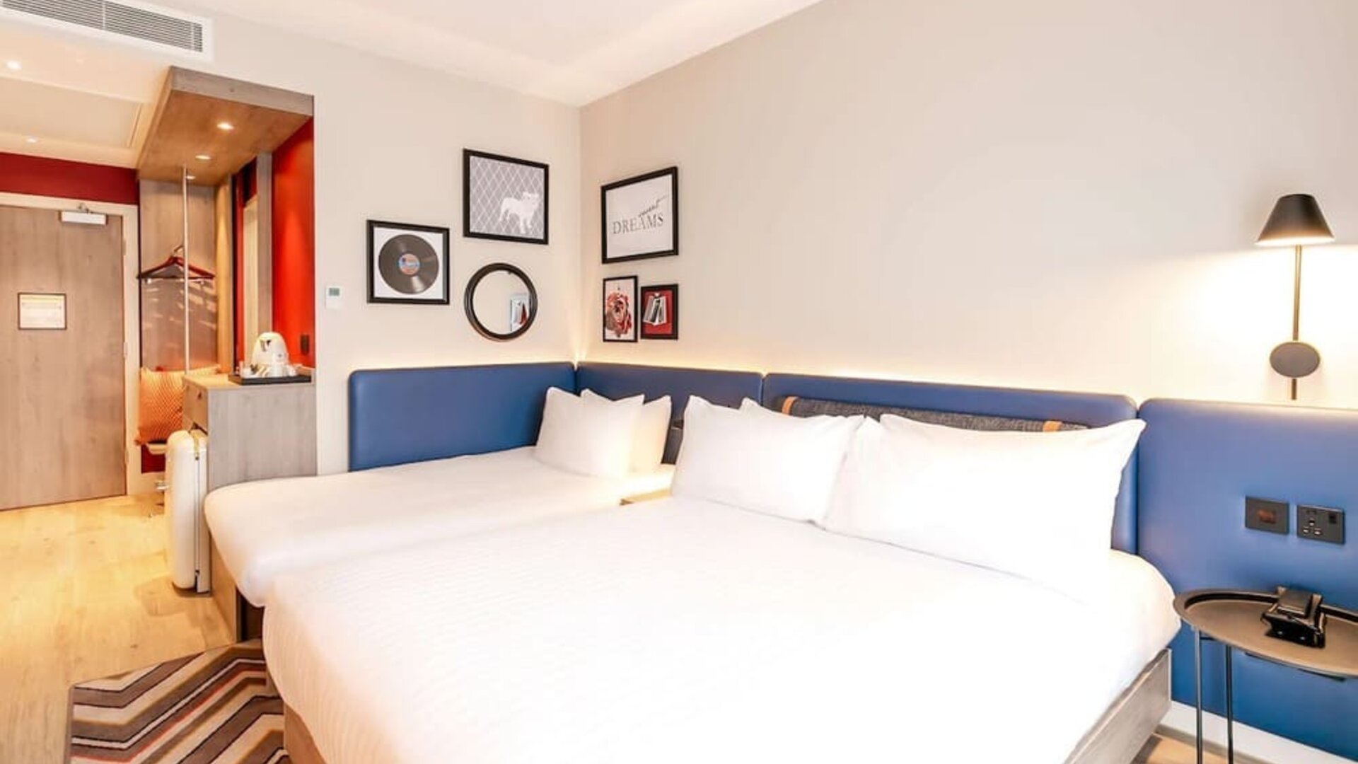 Contemporary hotel room with two beds dressed in white linens, long blue upholstered headboard, bedside lighting, framed artwork, patterned rug, tea and coffee station, and open wardrobe near the entrance.