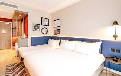 Contemporary hotel room with two beds dressed in white linens, long blue upholstered headboard, bedside lighting, framed artwork, patterned rug, tea and coffee station, and open wardrobe near the entrance.