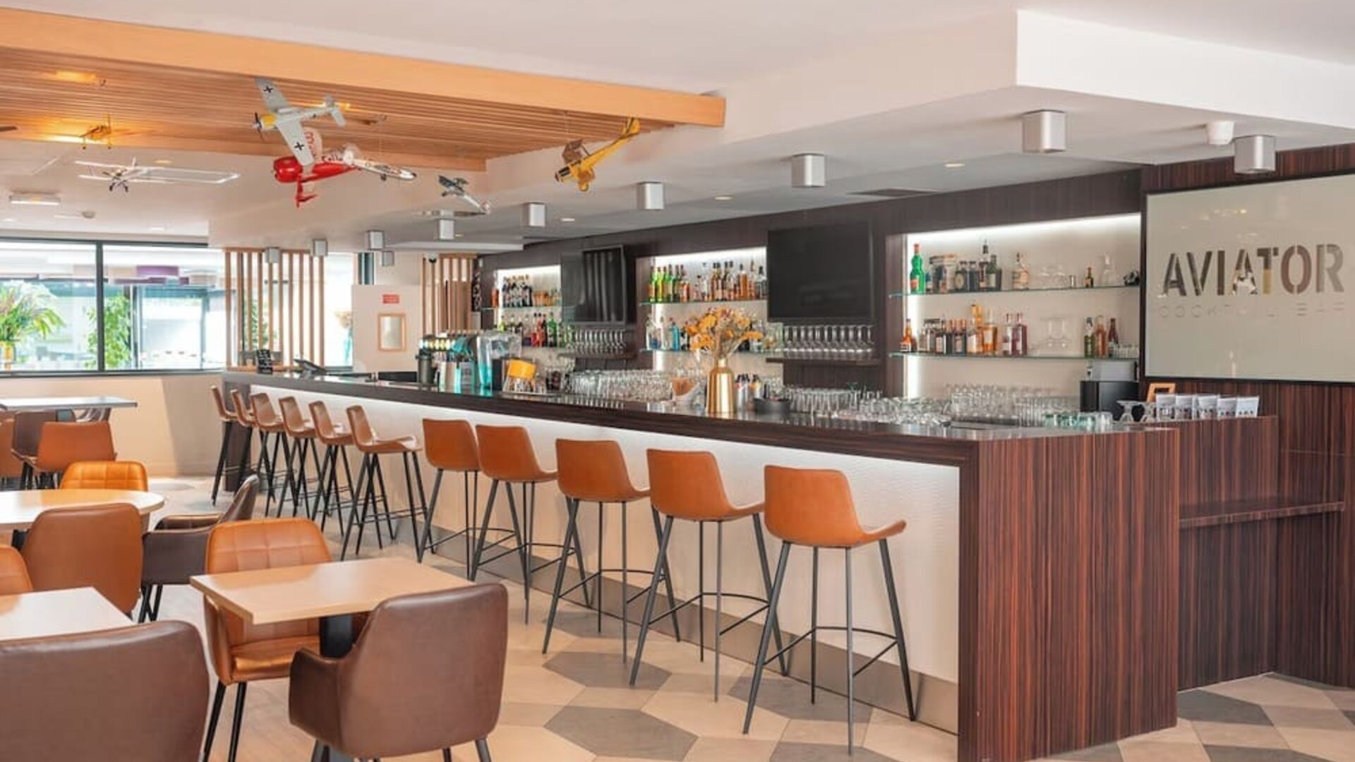 Hotel bar with a long counter, bar stools, lounge seating, airplane decorations, and illuminated shelving at Holiday Inn Toulouse Airport.
