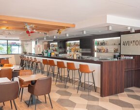 Hotel bar with a long counter, bar stools, lounge seating, airplane decorations, and illuminated shelving at Holiday Inn Toulouse Airport.