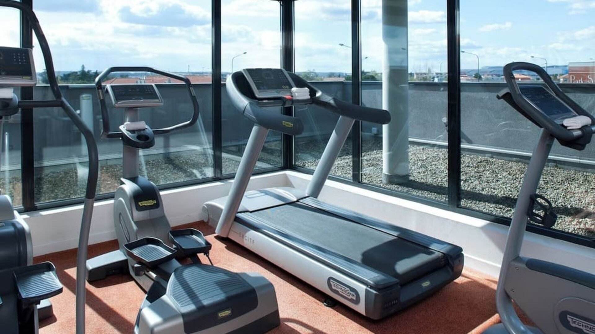 Hotel fitness room with treadmills, elliptical machines, and large windows overlooking the surrounding area at Holiday Inn Toulouse Airport.