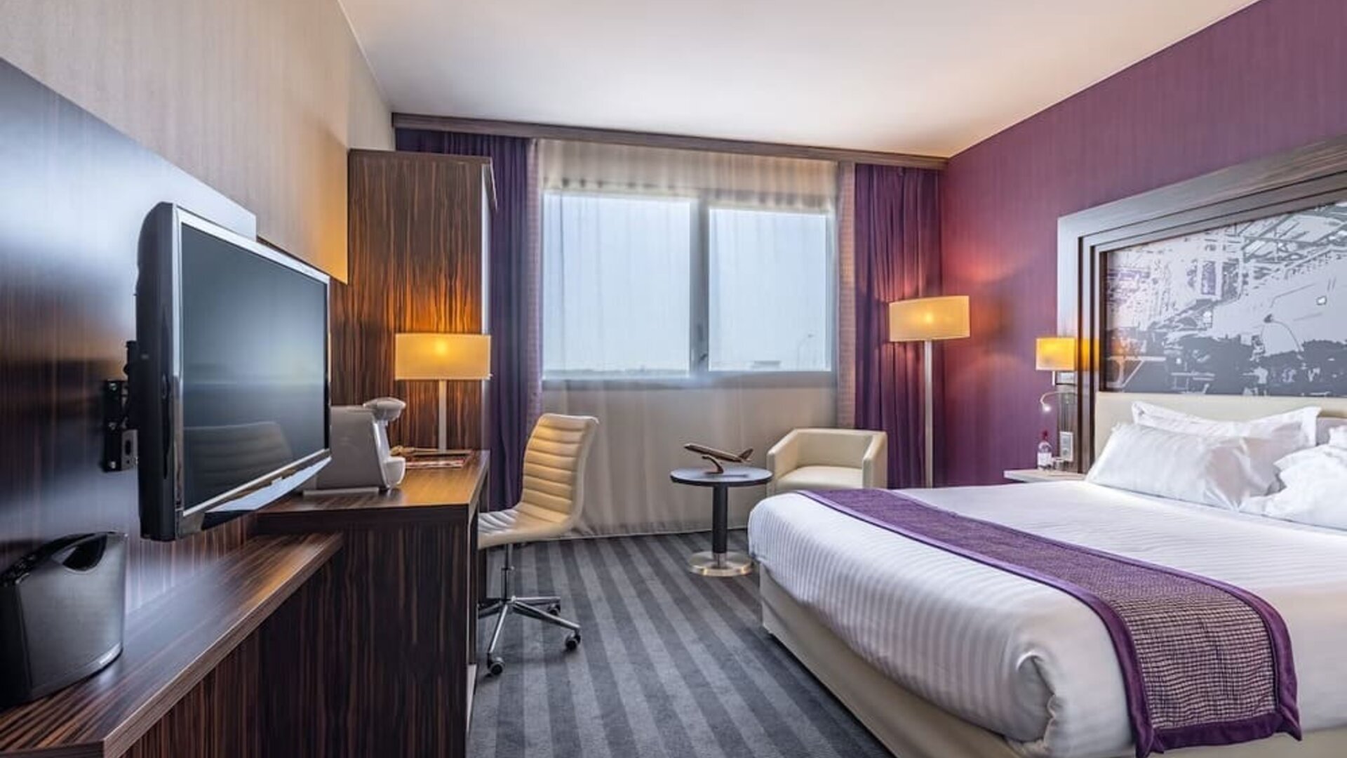 Guest room with a king bed, TV, work desk, armchair, wardrobe, and purple accent wall at Holiday Inn Toulouse Airport.