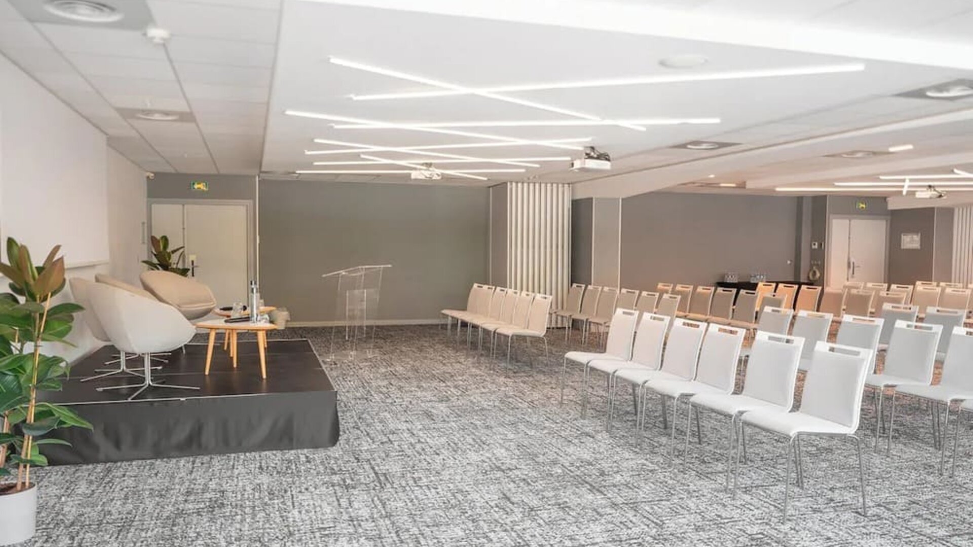 1. Large conference room with rows of white chairs, a presentation stage, podium, plants, and modern lighting at Holiday Inn Toulouse Airport.